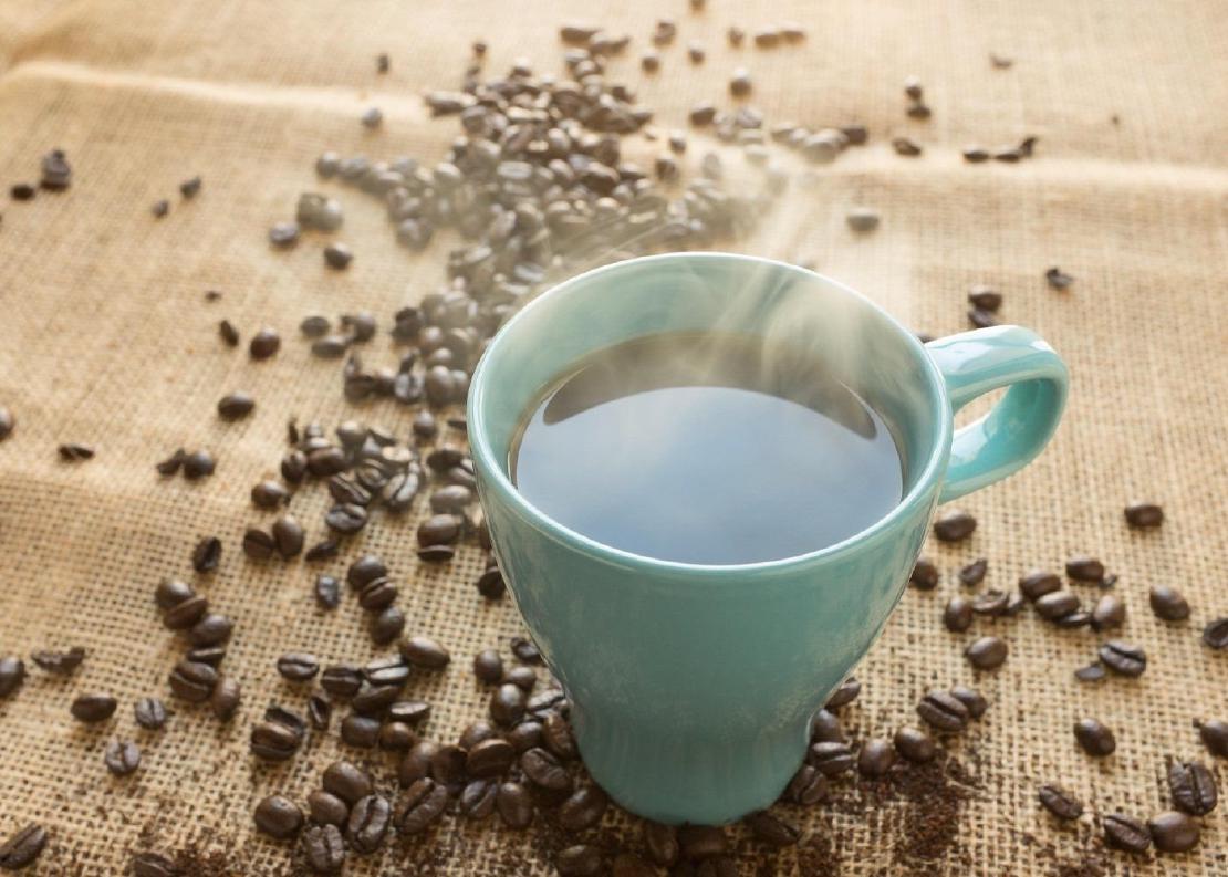 A cup of steaming coffee in a blue mug on a burlap surface surrounded by loose coffee beans.