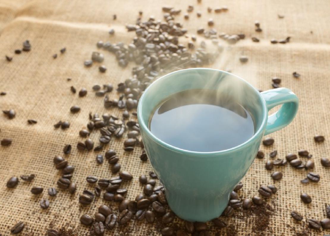 A cup of steaming coffee in a blue mug on a burlap surface surrounded by loose coffee beans.