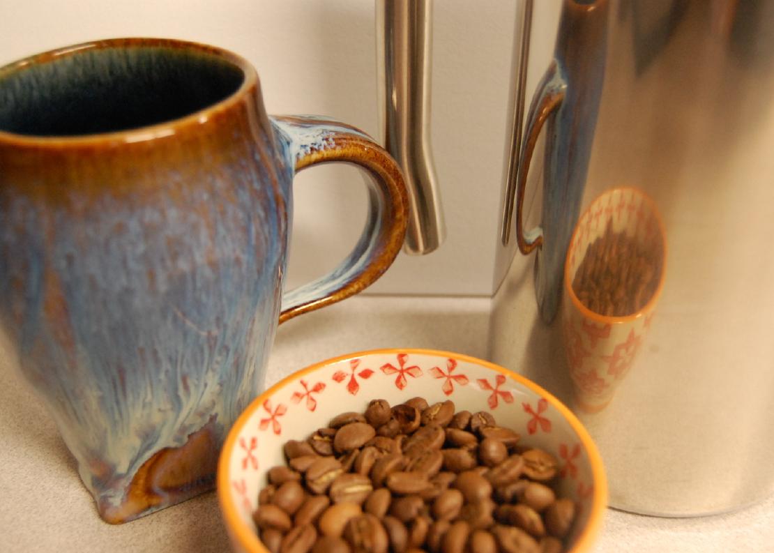 A close up image of a stainless French press, a ceramic cup, and a small bowl of unground coffee beans.