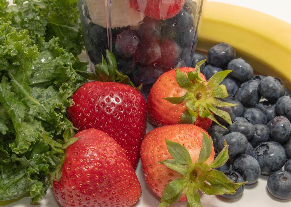 Strawberries, blueberries, a banana and some greens in front of a blender jar ready to make a smoothie.