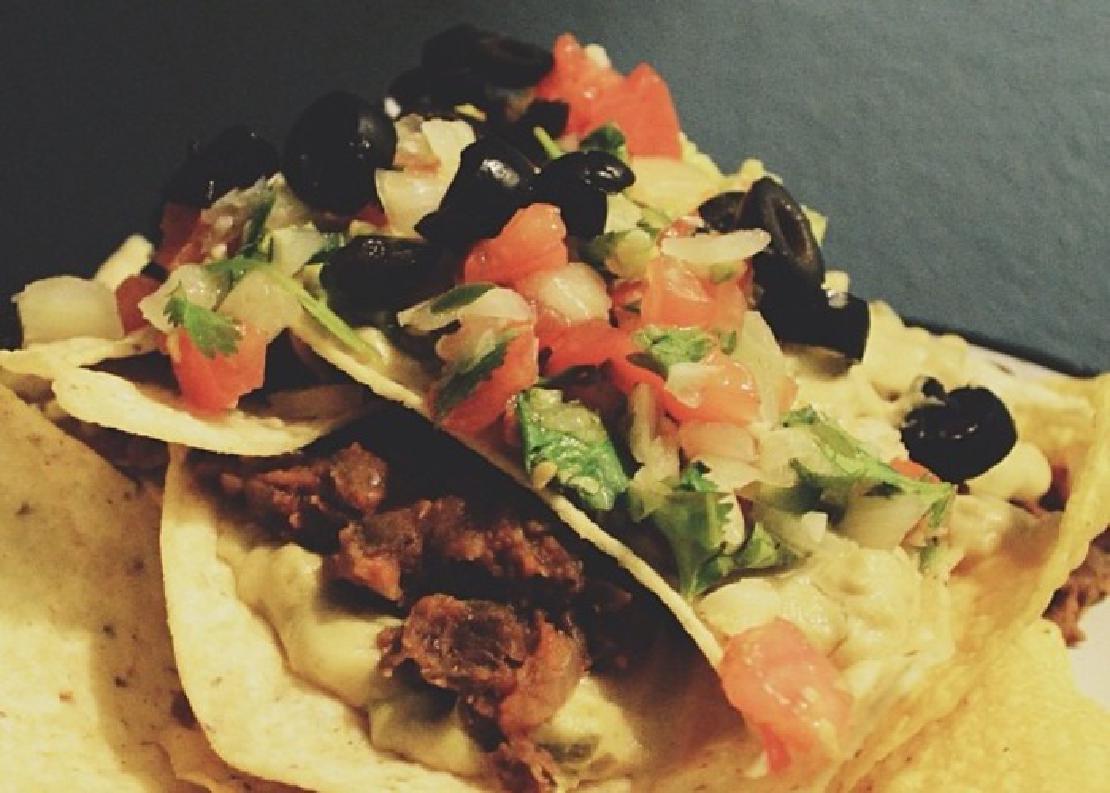 A close-up image of vegan nachos with tortilla chips, tomatoes, onions, plant-based meat and cheese, and black olives.