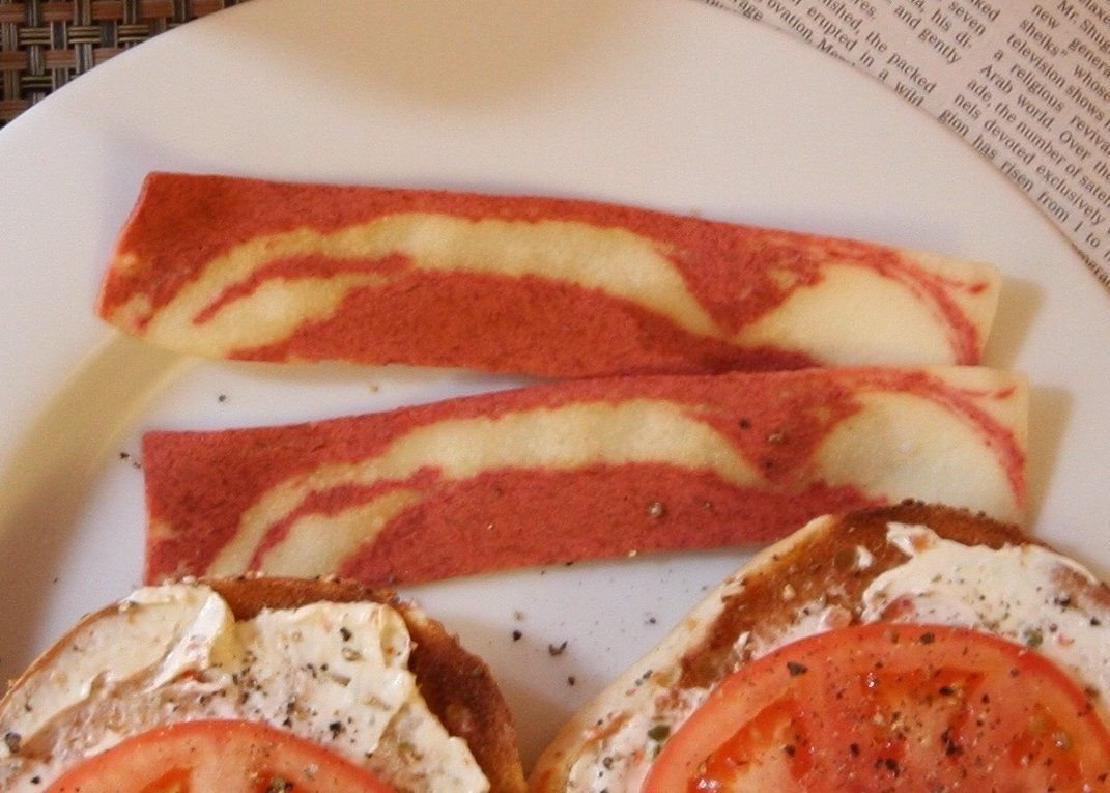 Two strips of Morningstar Farms Bacon on a plate above two bagel halves with cream spread and tomatoes on them.