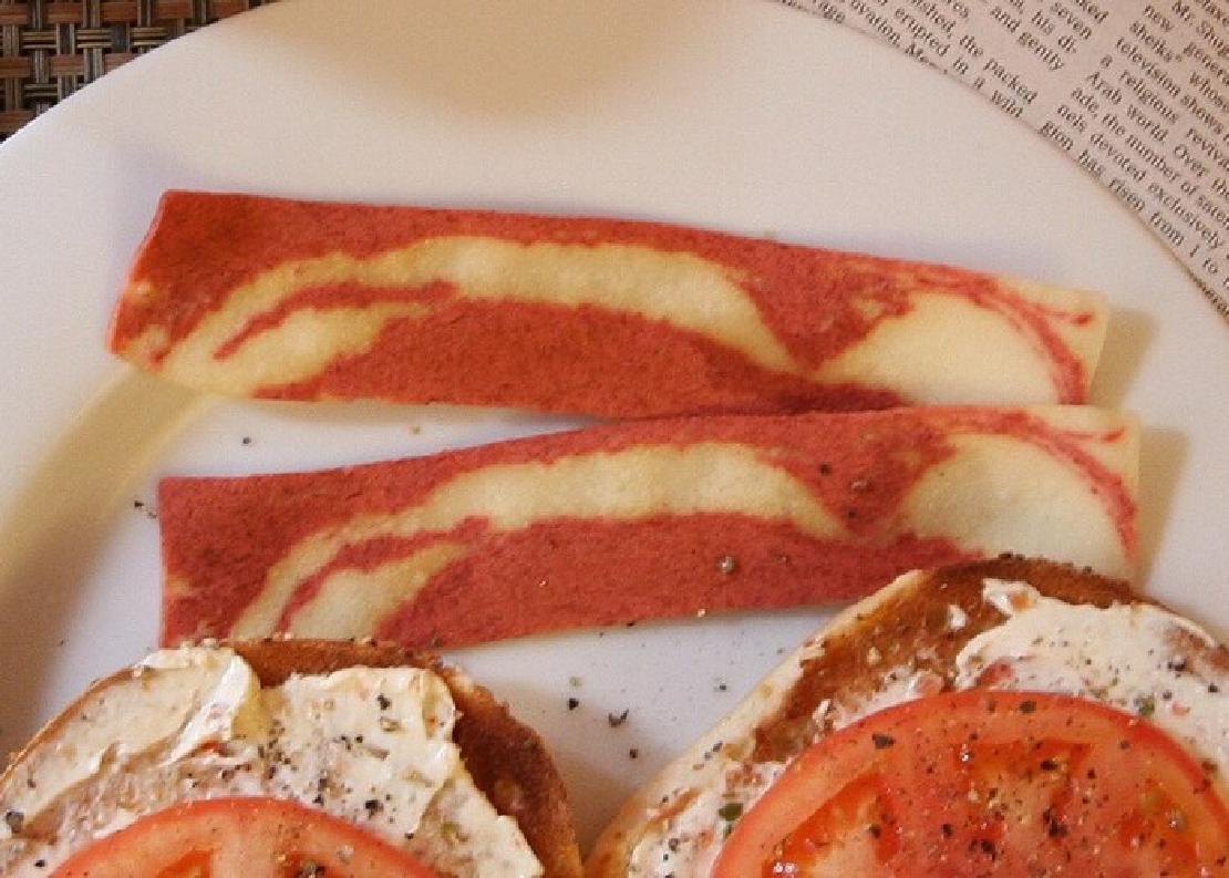 Two strips of Morningstar Farms Bacon on a plate above two bagel halves with cream spread and tomatoes on them.